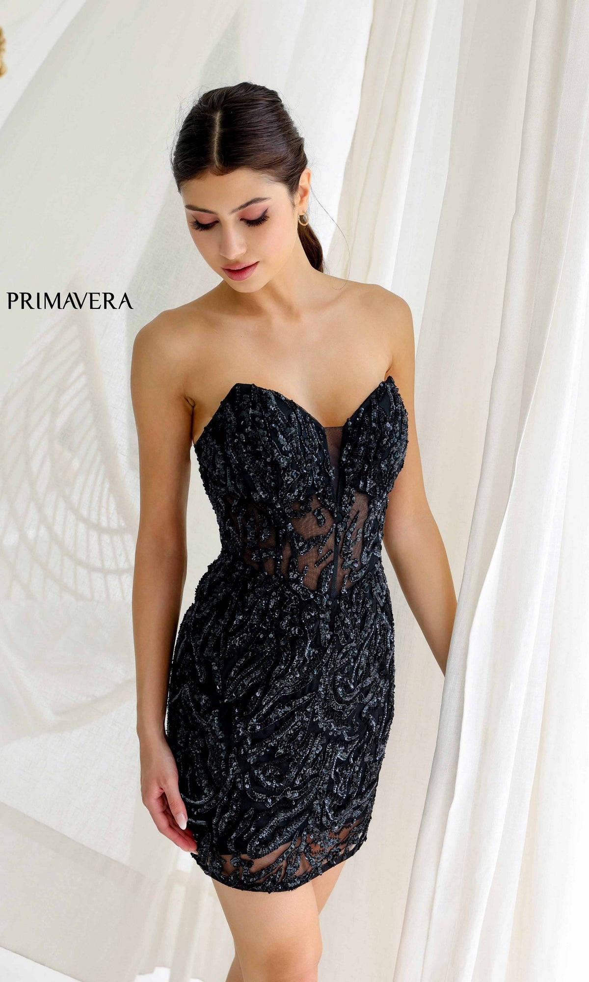 A woman stands by white curtains wearing the Short Homecoming Dress 4222 by Primavera—a strapless black mini dress featuring sequined beading, sheer details, and a corset bodice.